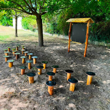 Plusieurs petits rondins servant de sièges pour enfants sont alignés sous des arbres, à l'ombre devant un tableau